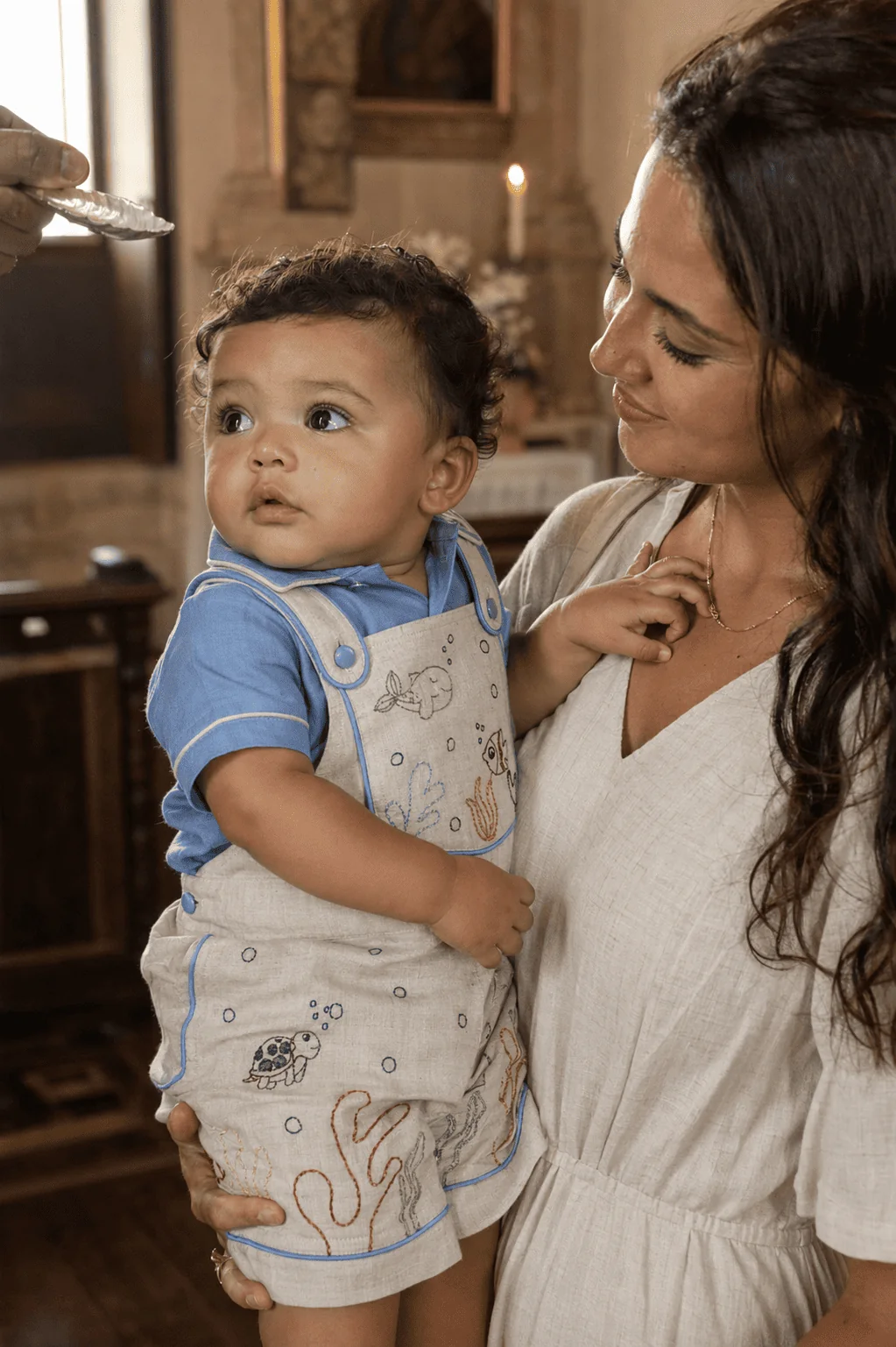 Mãe brasileira em vestido marfim segurando bebê de aproximadamente 8 meses no colo dentro de igreja católica histórica — bebê vestindo Jardineira Premium com camisa azul claro, pia batismal e velas ao fundo · cena de batizado formal, ambiente iluminado por luz natural atravessando vitrais